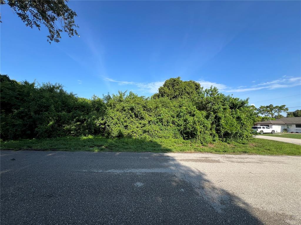 A vacant, overgrown residential lot sits along an asphalt street under a clear blue sky — 3239 MANHATTAN STREET, PORT CHARLOTTE, FL
