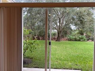 A view of a green backyard through a sliding glass door with vertical blinds — 6211 Arlington Way, Fort Pierce FL 34951, FORT PIERCE, FL