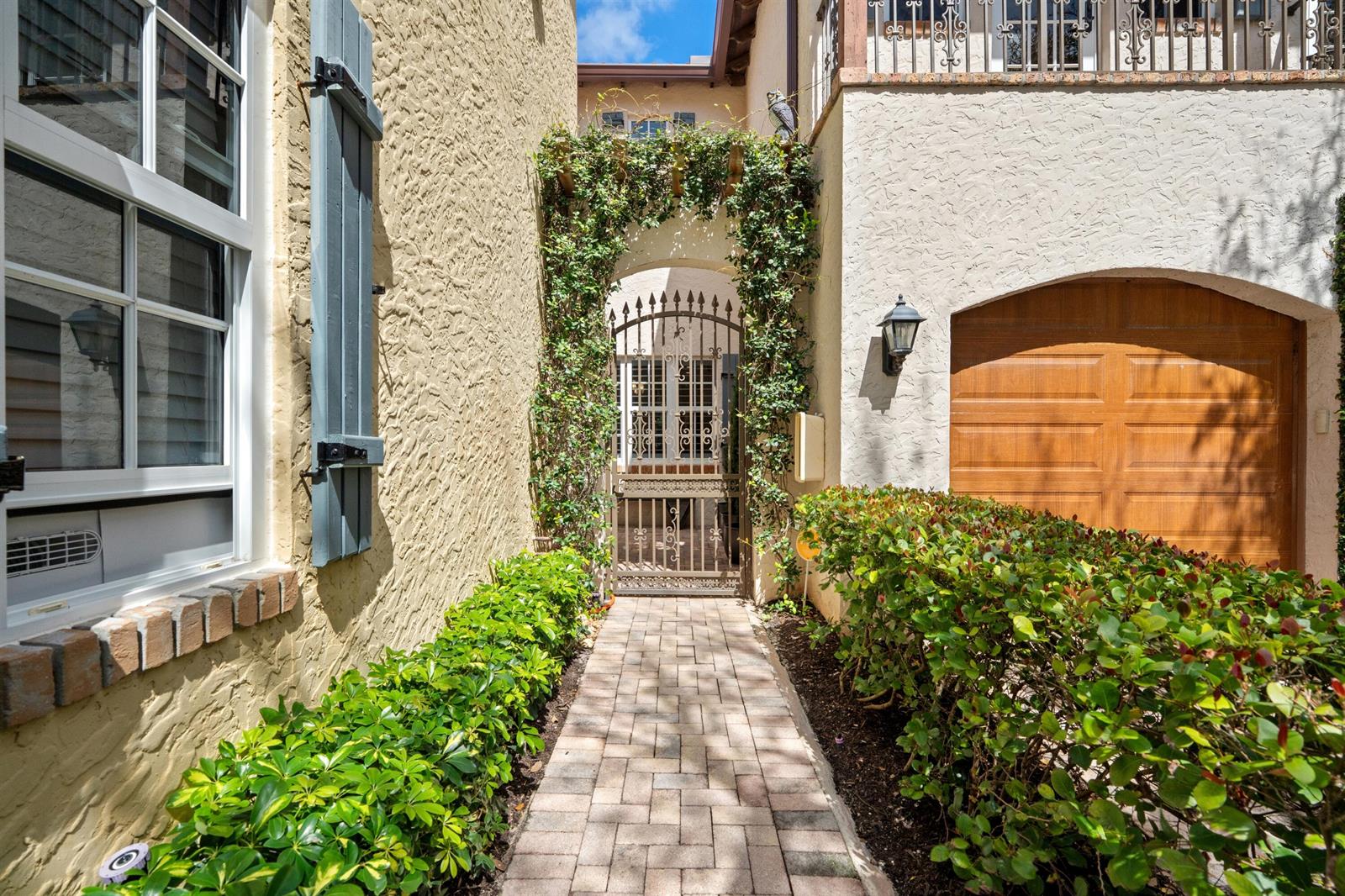 A paved walkway leads to an arched metal gate framed by climbing vines between two buildings — 1027 Coralina Lane, Delray Beach FL 33483, DELRAY BEACH, FL