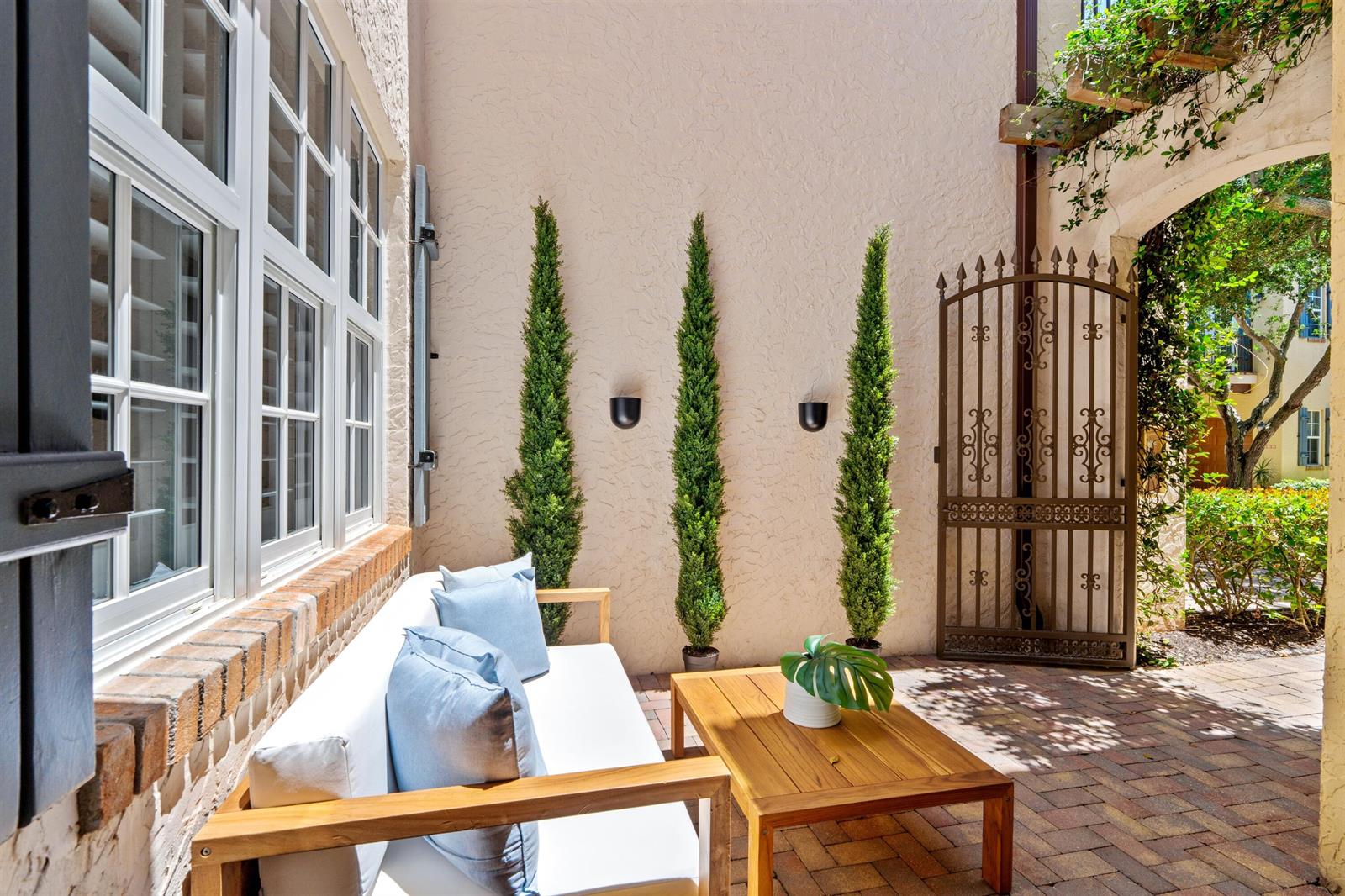A patio features a wooden sofa with blue pillows, a coffee table, and three cypress trees — 1027 Coralina Lane, Delray Beach FL 33483, DELRAY BEACH, FL