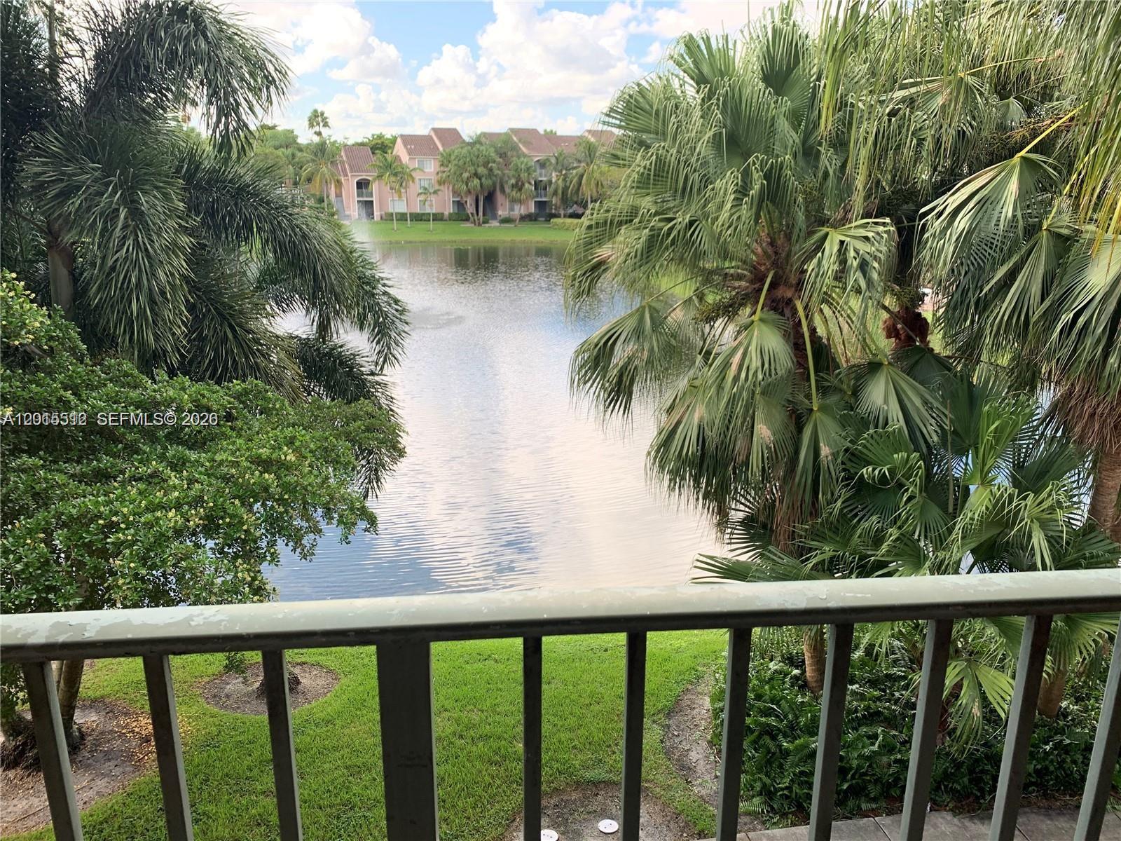 A balcony overlooks a lake with a fountain, palm trees, and apartment buildings in the distance — 12118 Saint Andrews Pl # 304, Miramar FL 33025, MIRAMAR, FL