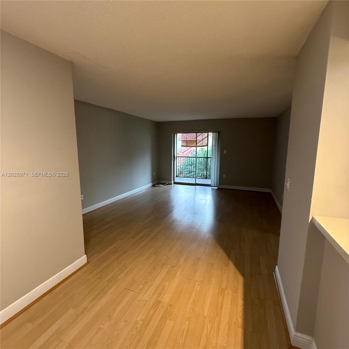 An empty living room features light wood-look flooring, gray walls, and a sliding glass door — POMPANO BEACH, FL