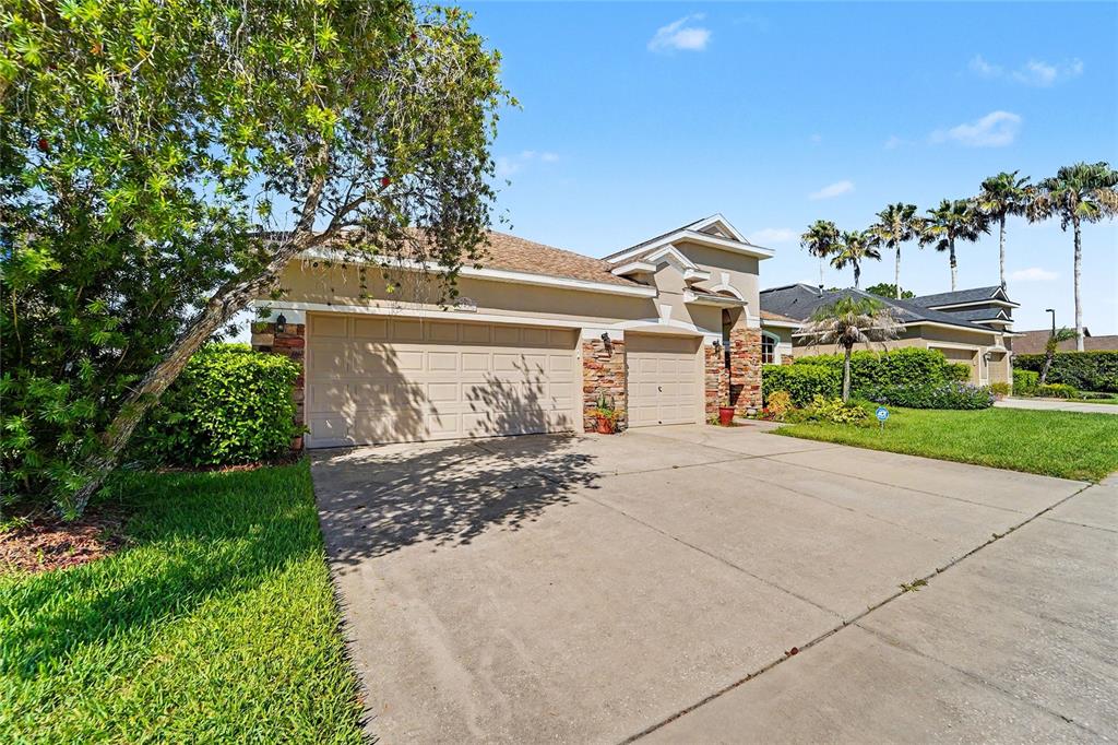A single-story house with a three-car garage and stone accents sits under a blue sky — 27236 HOLLYBROOK TRAIL, WESLEY CHAPEL, FL
