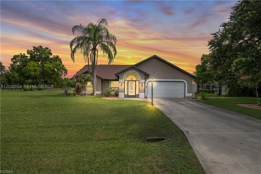 A single-story house with a two-car garage and a palm tree sits on a lawn — 538 Whispering Wind Bnd, Lehigh Acres FL 33974, LEHIGH ACRES, FL