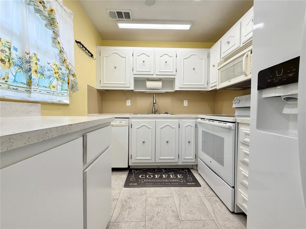 A kitchen features white cabinetry, white appliances, a light-colored countertop, and a sunflower-patterned window curtain — 11817 BAYONET LANE, NEW PORT RICHEY, FL