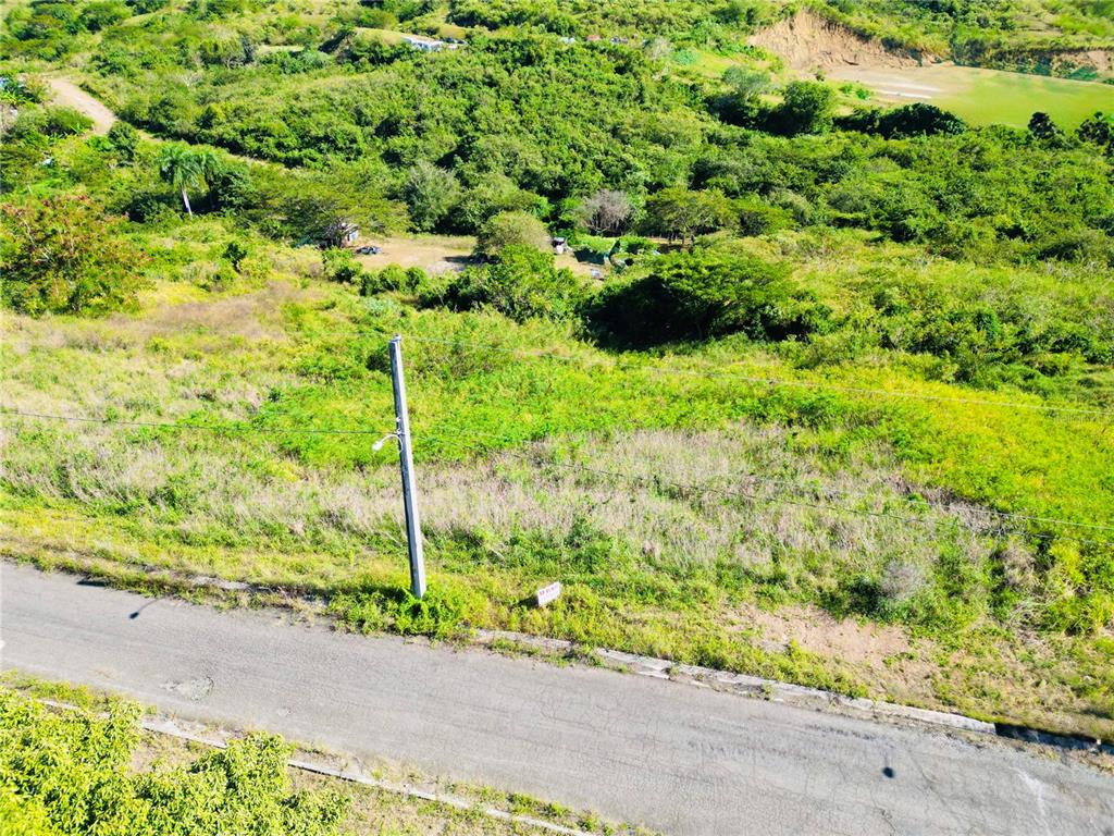 An aerial view shows a paved road bordering a grassy, undeveloped plot of land — URB.VISTAS DEL RODADERO, YAUCO, PR