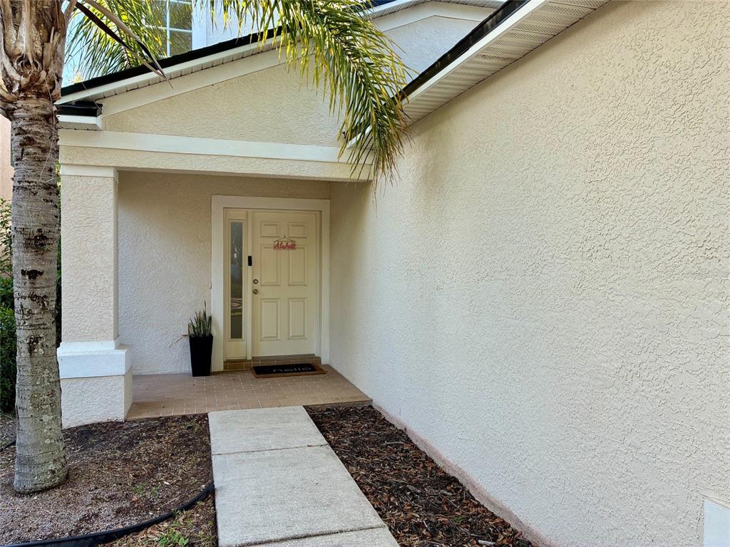 A concrete walkway leads to the covered entryway of a house with beige stucco walls — 8026 CARRIAGE POINTE DRIVE, GIBSONTON, FL