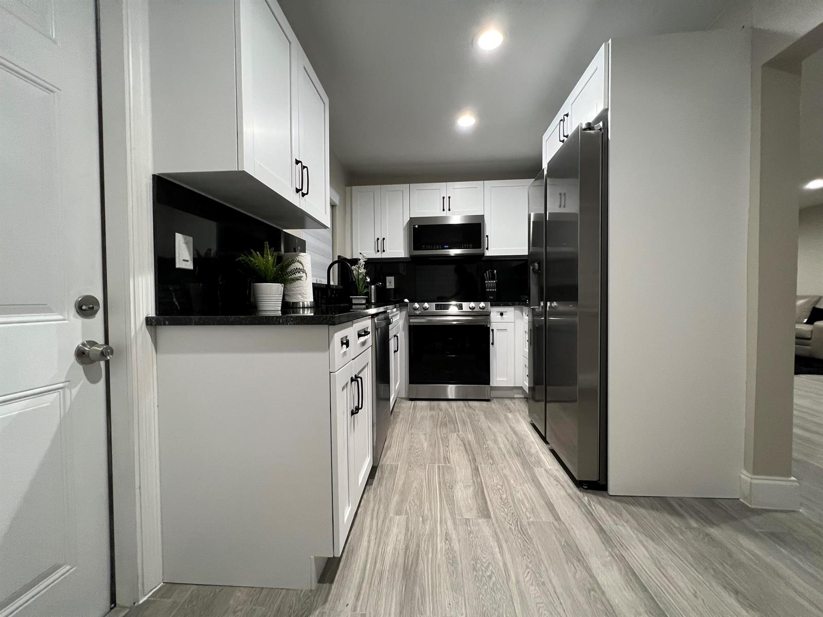 A galley kitchen features white cabinets, black countertops, stainless steel appliances, and wood-look flooring — 601 NW 2nd Street, Delray Beach FL 33444, DELRAY BEACH, FL