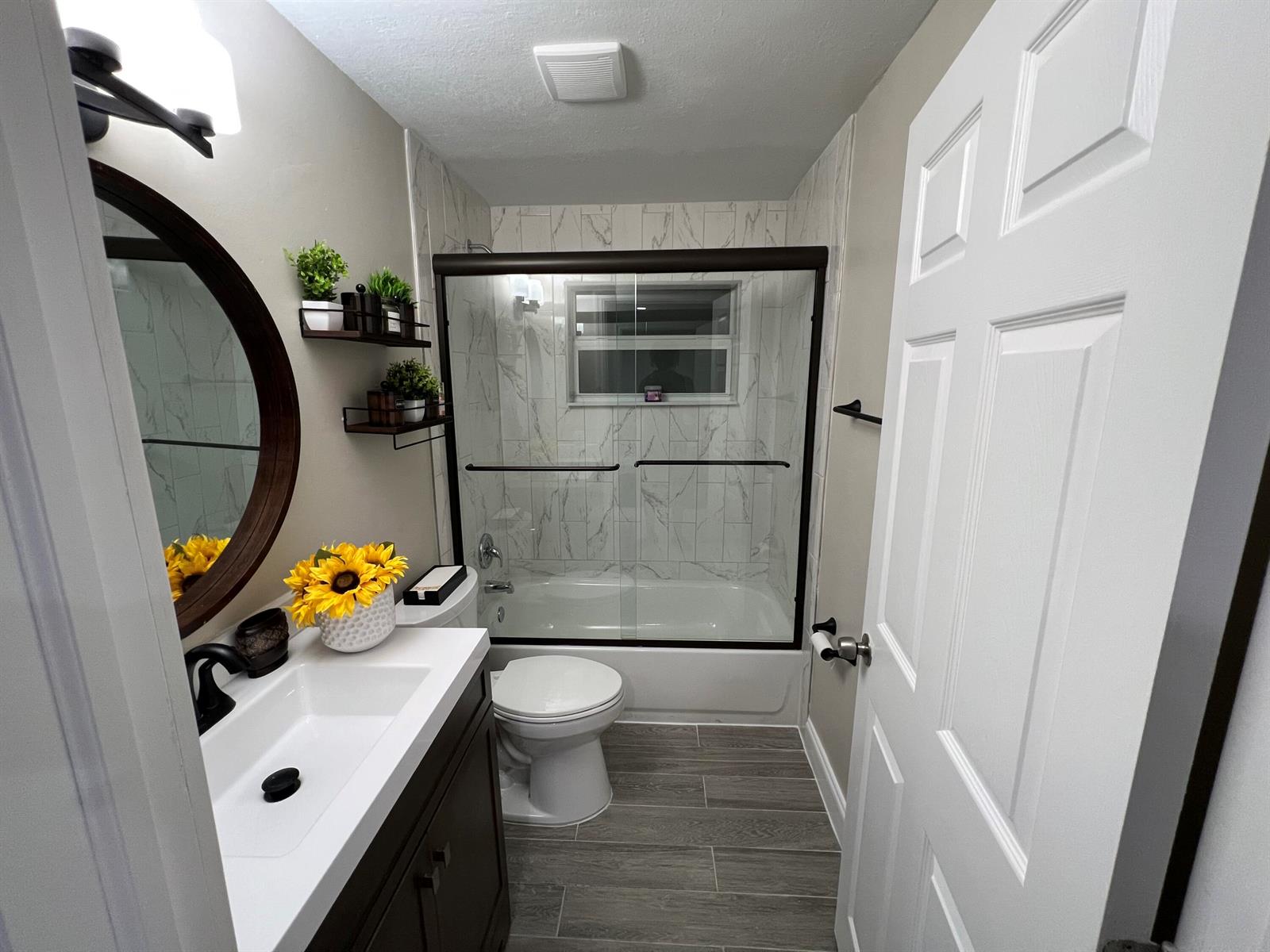 A bathroom features a vanity with a round mirror, a toilet, and a tiled tub-shower — 601 NW 2nd Street, Delray Beach FL 33444, DELRAY BEACH, FL
