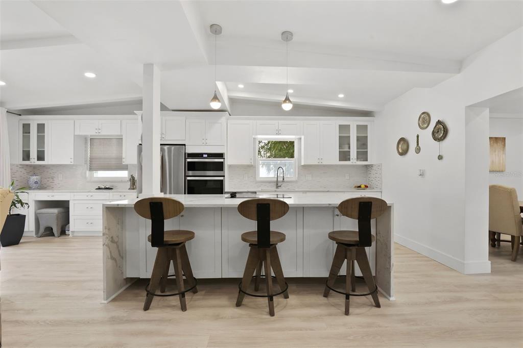 A modern white kitchen features a large island with three wooden stools and pendant lighting — 25 SANDPIPER ROAD, TAMPA, FL