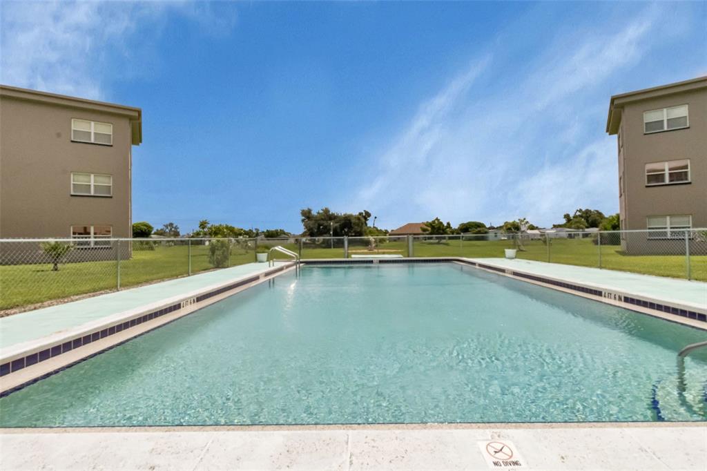 A rectangular swimming pool sits between two multi-story apartment buildings under a blue sky — 3100 HARBOR BOULEVARD UNIT 203, PORT CHARLOTTE, FL