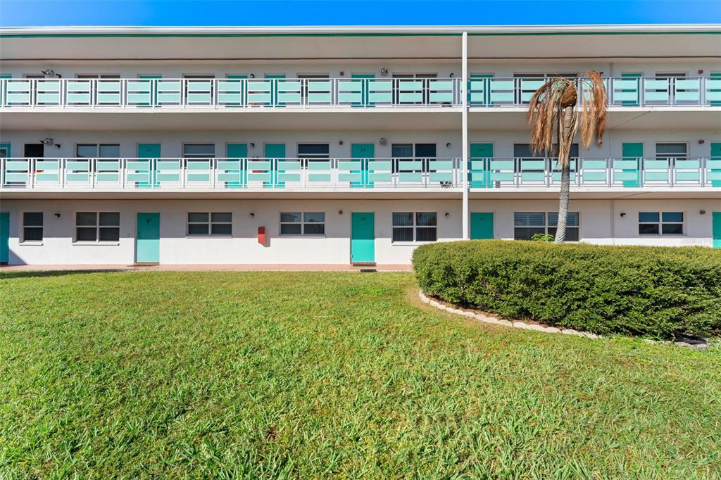 A three-story apartment building features teal doors, white railings, and a manicured lawn — 1950 59TH AVENUE N UNIT 114, ST PETERSBURG, FL