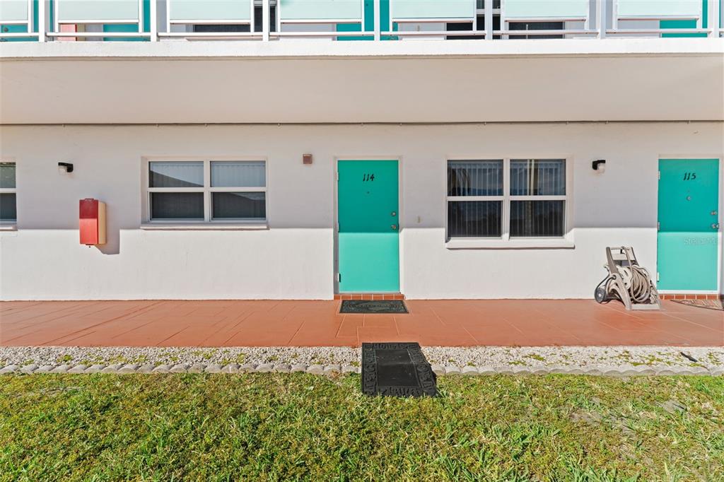 Exterior view of a ground-level apartment building with teal doors numbered 114 and 115 — 1950 59TH AVENUE N UNIT 114, ST PETERSBURG, FL