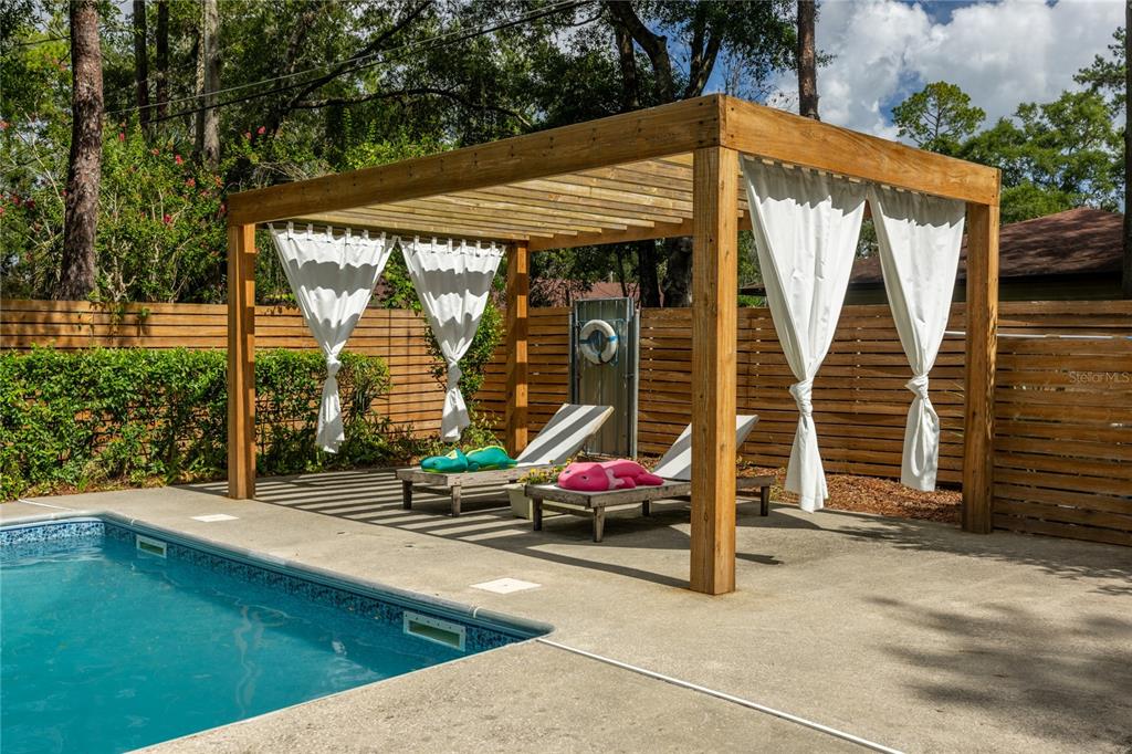 A wooden pergola with white curtains stands over two lounge chairs beside a swimming pool — 4204 NW 19TH PLACE, GAINESVILLE, FL