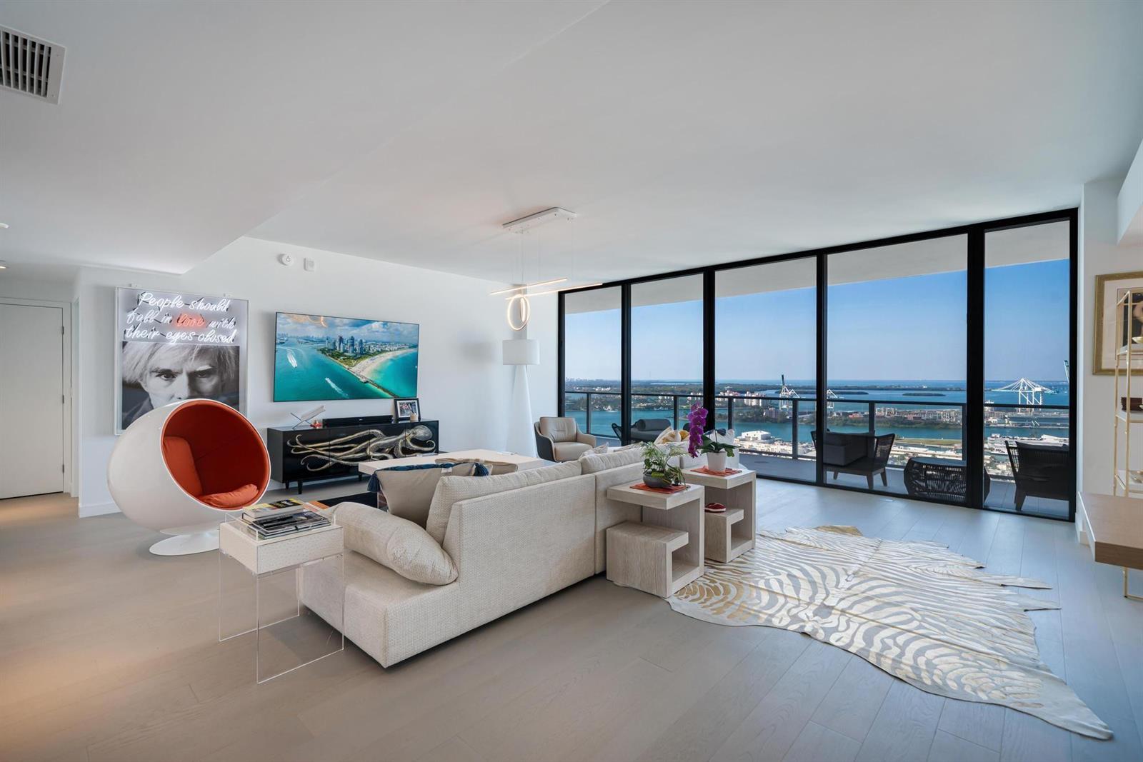 A modern living room with floor-to-ceiling windows overlooking a city skyline and harbor — 500 Alton # 2702, Miami Beach FL 33139, MIAMI BEACH, FL