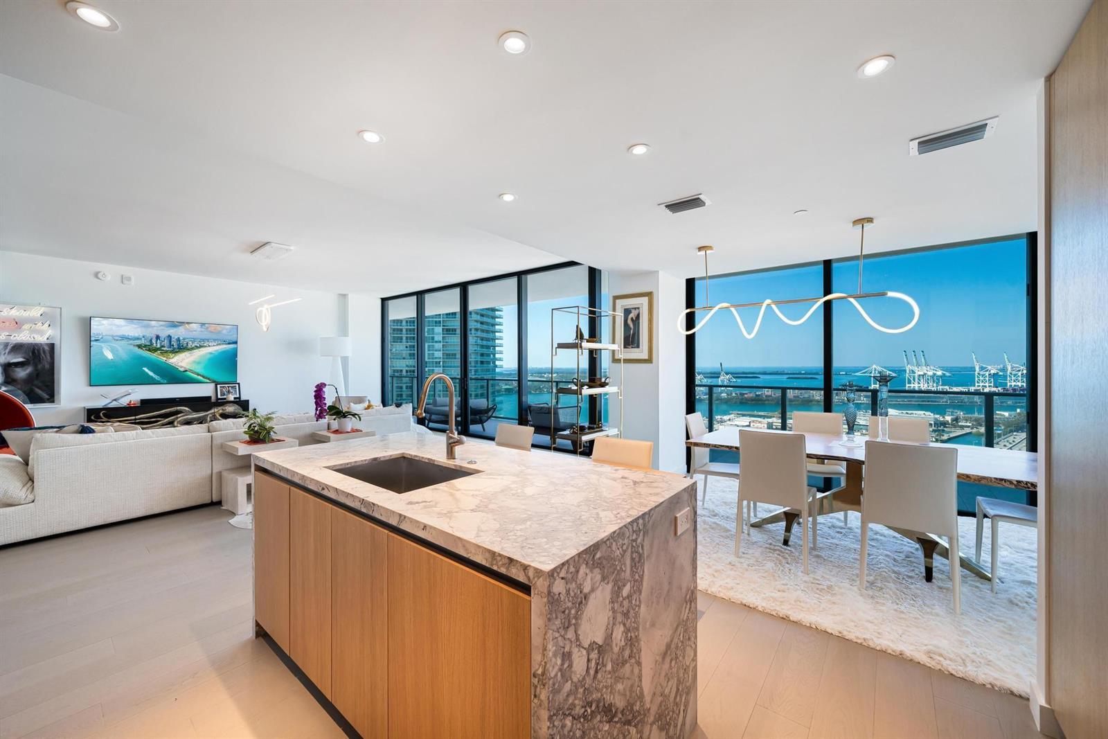 A modern open-concept kitchen and dining area with marble island, wood cabinetry, and harbor views — 500 Alton # 2702, Miami Beach FL 33139, MIAMI BEACH, FL