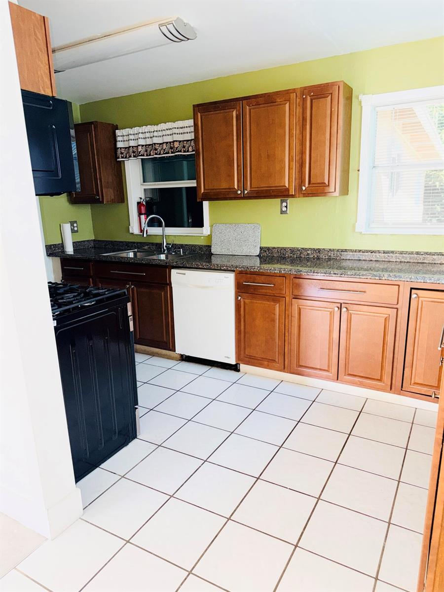 A kitchen features wooden cabinets, granite countertops, a white dishwasher, and white tiled flooring — 5755 Winfield Boulevard, Margate FL 33063, MARGATE, FL
