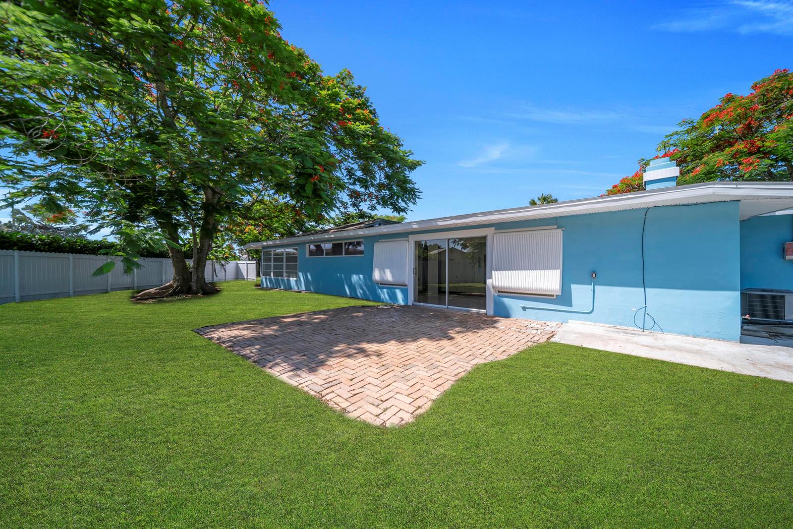 A blue house exterior features a brick patio, a large tree, and a fenced yard — 139 E Lake Drive, Lehigh Acres FL 33936, LEHIGH ACRES, FL