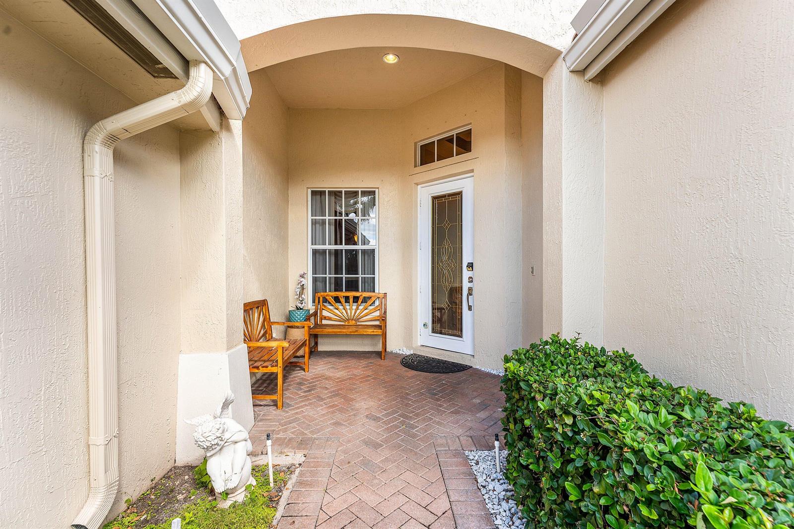 A covered entryway features a brick patio, two wooden benches, and a white front door — 6529 Jog Estates Lane, Boynton Beach FL 33437, BOYNTON BEACH, FL