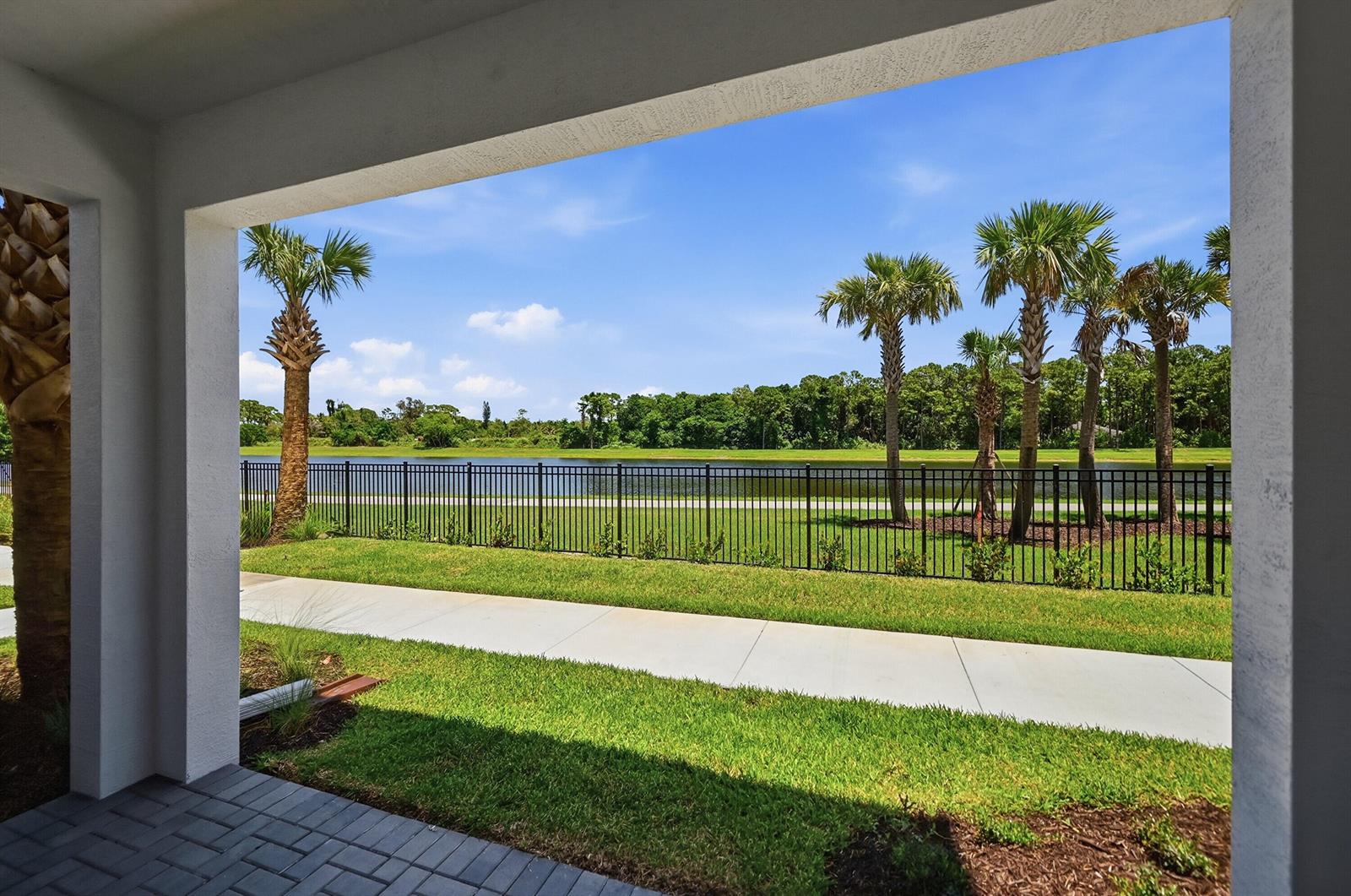 A covered patio overlooks a grassy lawn, a black metal fence, a pond, and palm trees — 4356 Brookgreen Square, Westlake FL 33470, WESTLAKE, FL