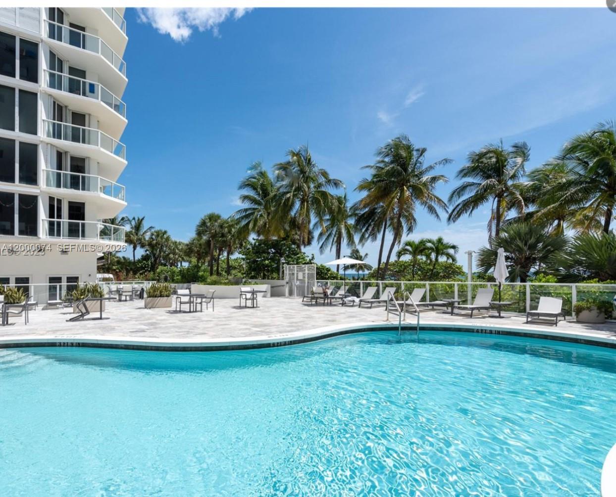 A swimming pool with a patio area, lounge chairs, and palm trees beside a tall building — 8925 Collins Ave # 2J, Surfside FL 33154, SURFSIDE, FL