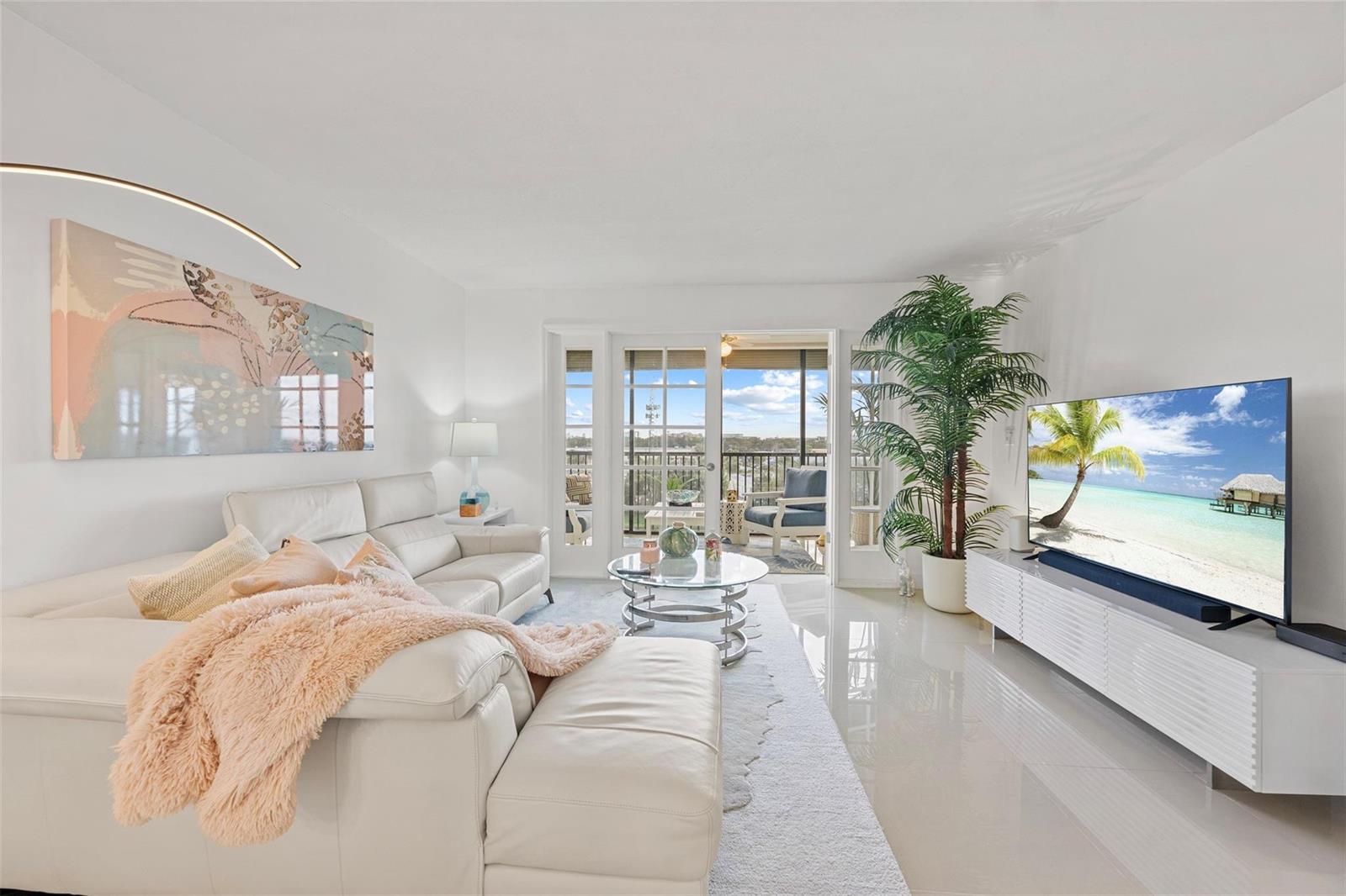 A bright living room features white leather furniture, a glass coffee table, and a balcony view — 2202 S Cypress Bend Drive # 606, Pompano Beach FL 33069, POMPANO BEACH, FL