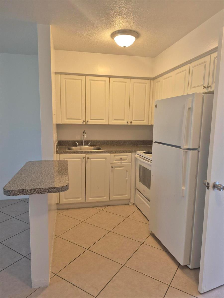 A small kitchen features white cabinets, a white refrigerator, a sink, and tiled flooring — 1960 NE 48 Street # 9, Pompano Beach FL 33064, POMPANO BEACH, FL