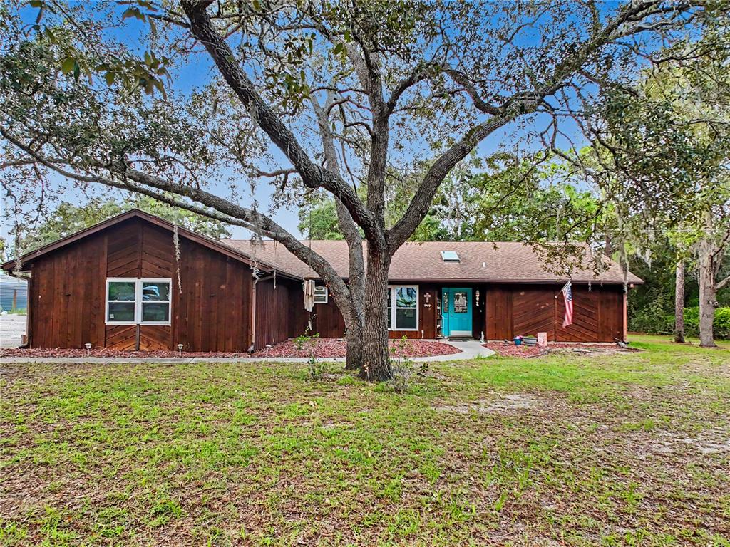A single-story house with dark wood siding, a bright blue door, and a large tree — 5253 W ROCHELLE STREET, HOMOSASSA, FL