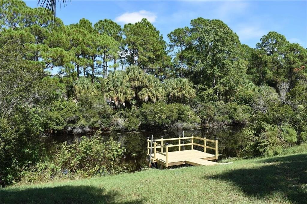 A wooden dock sits on a small pond surrounded by dense trees and green grass — 2984 TUSKET AVENUE, NORTH PORT, FL