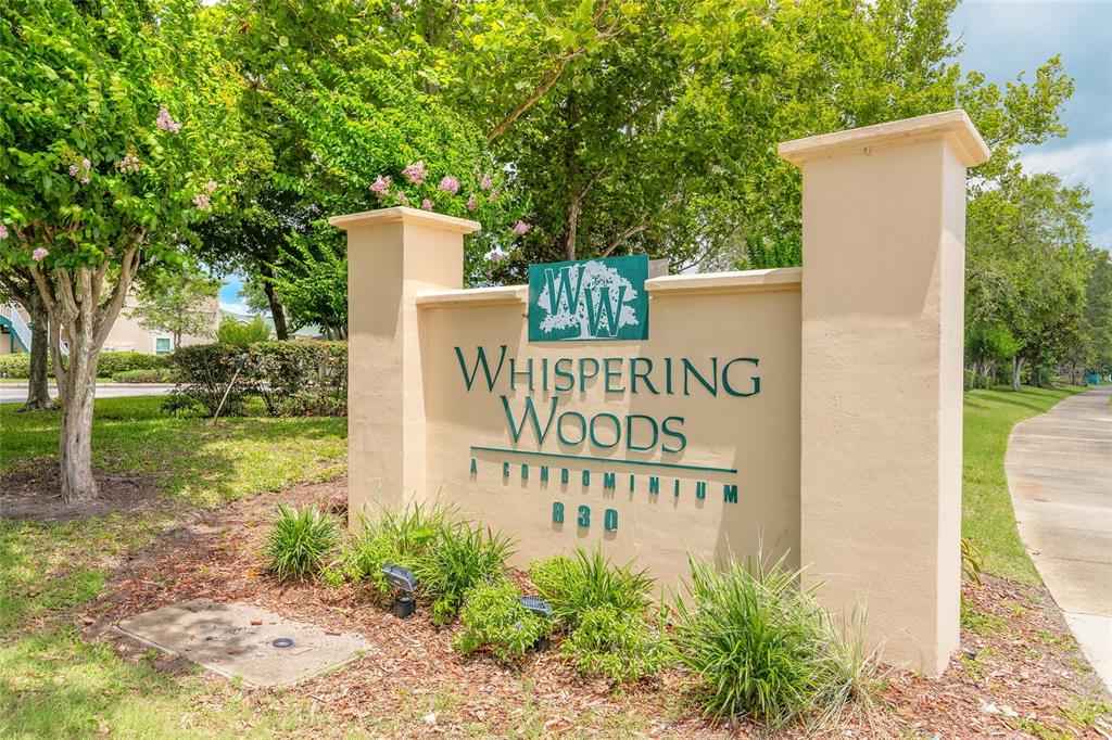 A tan stucco monument sign for Whispering Woods Condominium stands in a landscaped outdoor area — 830 AIRPORT ROAD UNIT 105, PORT ORANGE, FL