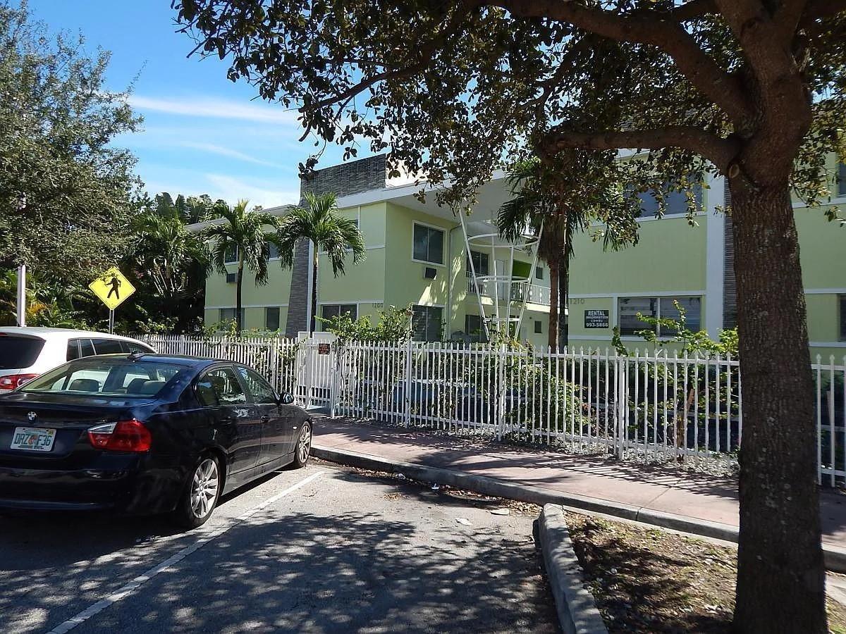 A light green two-story apartment building is viewed from the street behind a white fence — 1200 Marseille Drive, Miami Beach FL 33141, MIAMI BEACH, FL