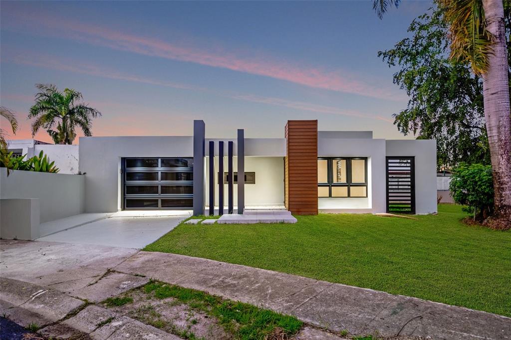 A modern single-story house features a flat roof, wood-paneled accent wall, and a glass garage door — RAMON MARIN ST. LEVITTOWN LAKES UNIT EA-34, TOA BAJA, PR
