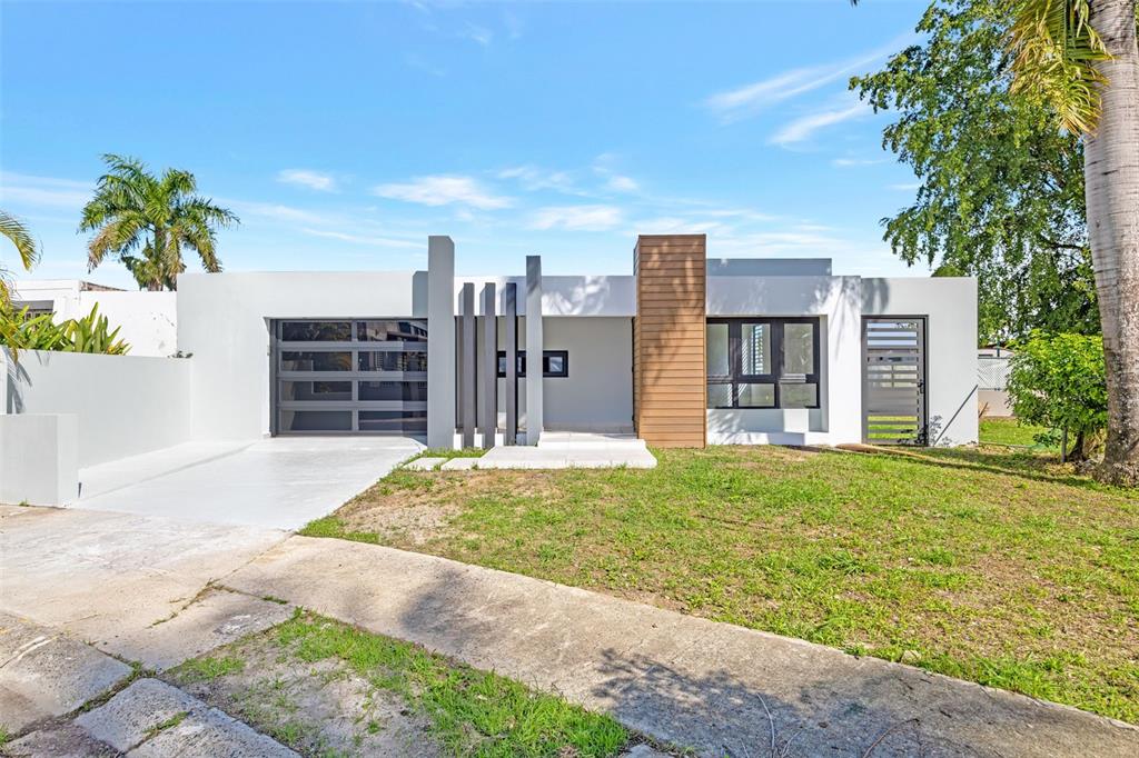 A modern, single-story house with a white exterior, wood-paneled accent, and a gray garage door — RAMON MARIN ST. LEVITTOWN LAKES UNIT EA-34, TOA BAJA, PR