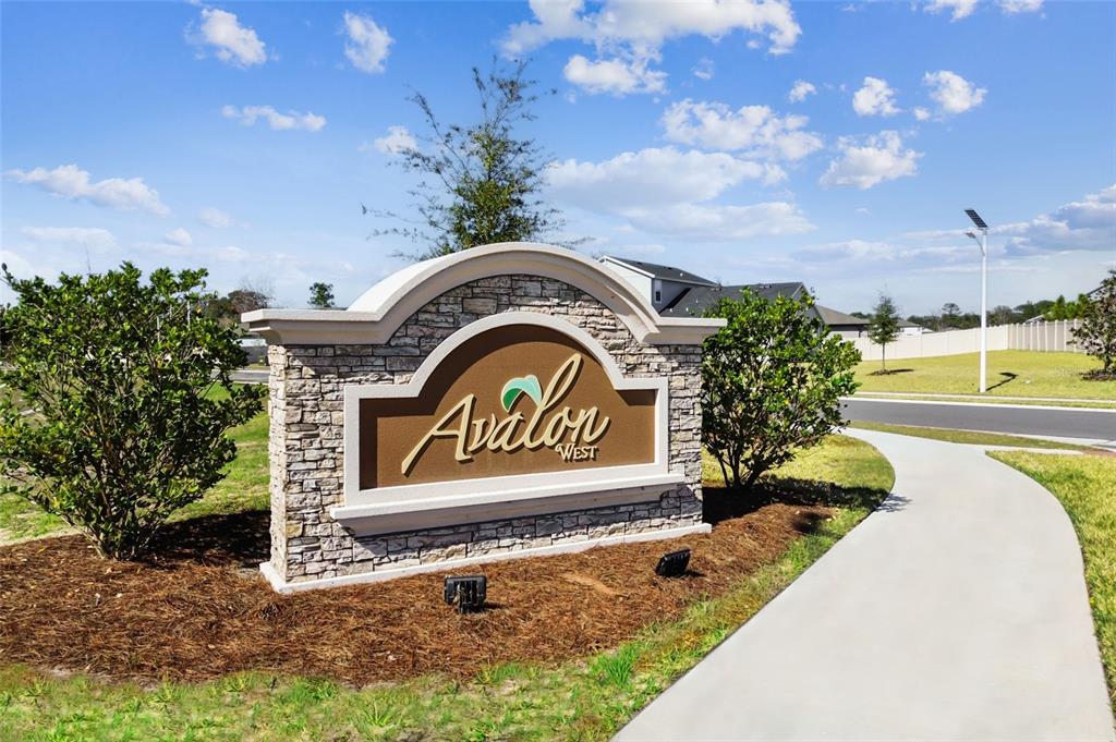 A stone monument sign for Avalon West stands in a landscaped yard near a sidewalk — 105 NORTHBANK LANE, SPRING HILL, FL