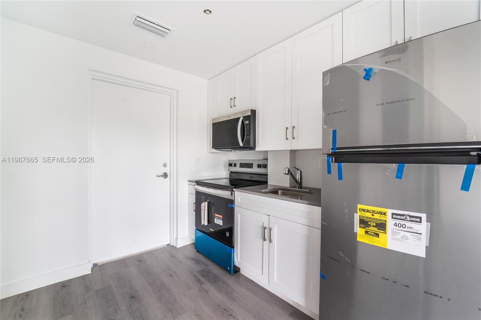 A small kitchen features white cabinets, stainless steel appliances, and gray wood-look flooring — 1165 NW 8th Ave # 101, Miami FL 33136, MIAMI, FL