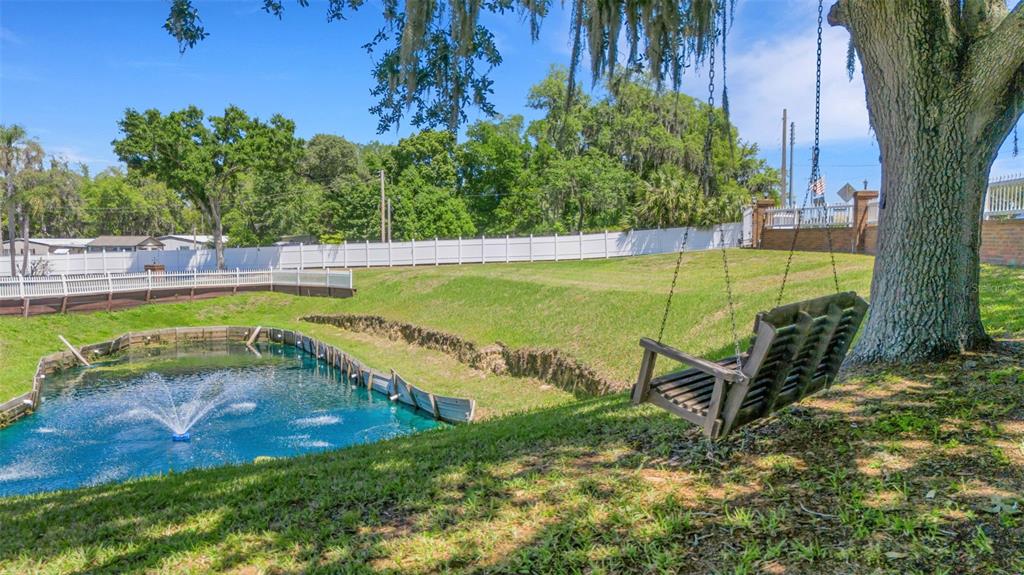A wooden porch swing hangs from a large tree overlooking a small pond with fountains — 5905 MAGNOLIA LANE, LAKELAND, FL