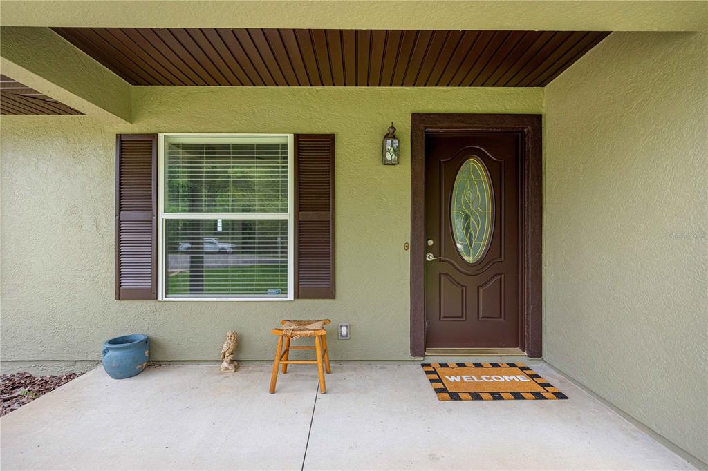 A covered concrete porch features a brown front door, a window with shutters, and decor — 80 PECAN RUN COURSE, OCALA, FL