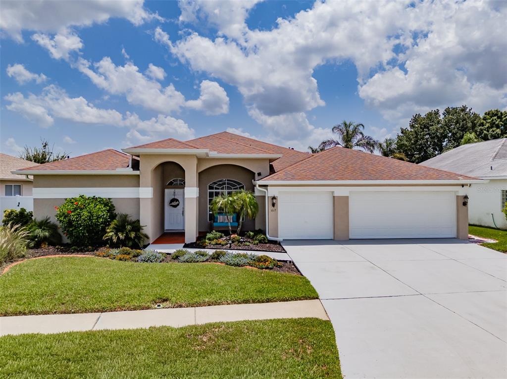 A single-story house with a tan stucco exterior, a two-car garage, and a landscaped front yard — 1617 VISTOSO LANE, SUN CITY CENTER, FL