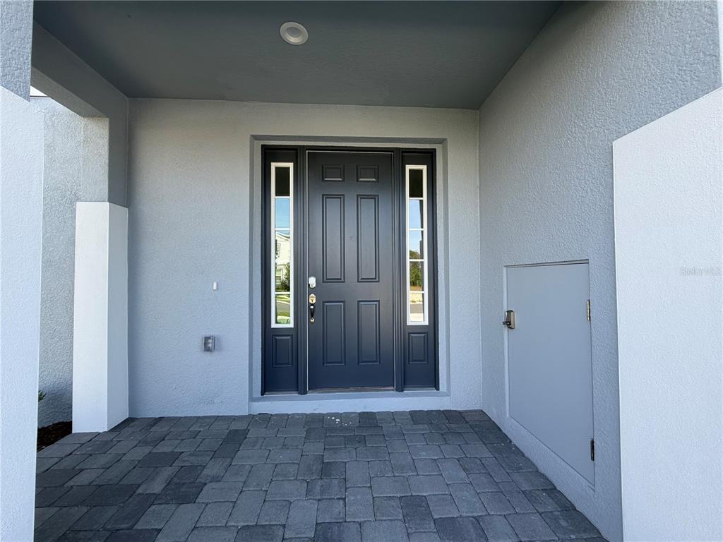 A gray front door with sidelights sits under a covered porch with paver flooring — 756 FARFIELDS STREET, DELAND, FL