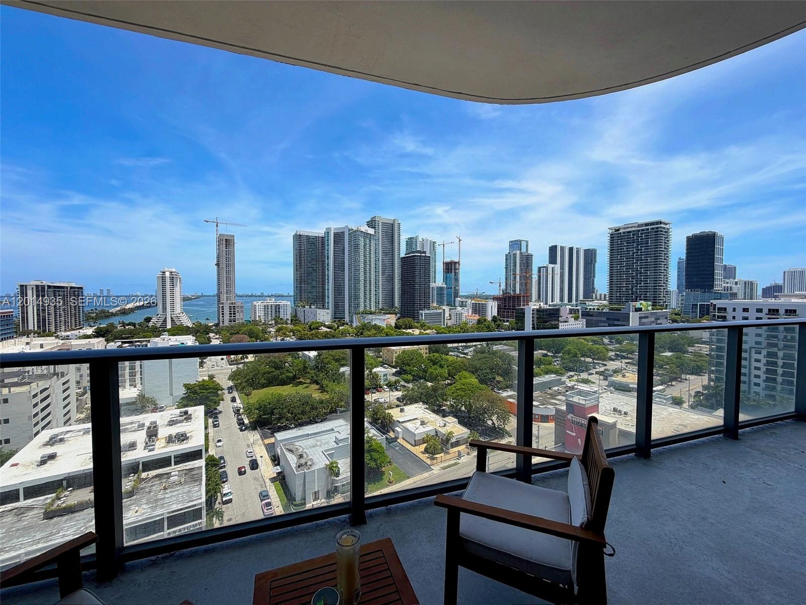 A balcony with a wooden chair and table overlooks a city skyline and water — 121 NE 34th St # 1605, Miami FL 33137, MIAMI, FL
