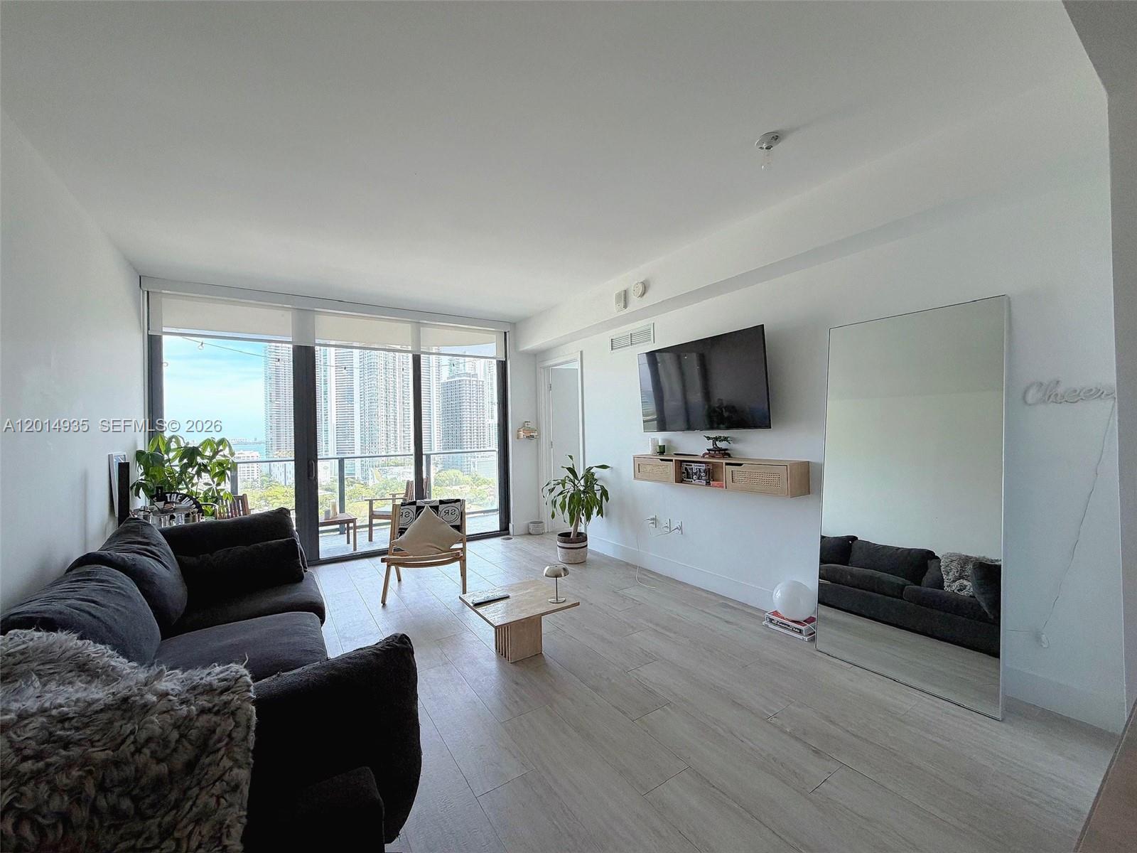 A modern living room features a dark sofa, a wall-mounted television, and floor-to-ceiling windows — 121 NE 34th St # 1605, Miami FL 33137, MIAMI, FL