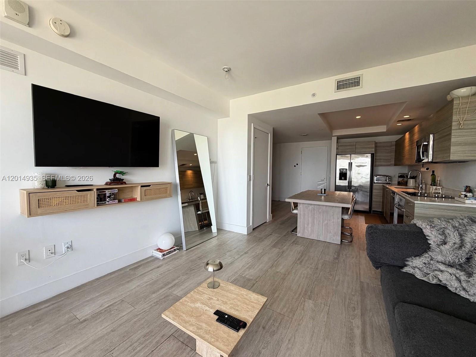 A living room with light wood flooring, a wall-mounted television, and an open-concept kitchen — 121 NE 34th St # 1605, Miami FL 33137, MIAMI, FL