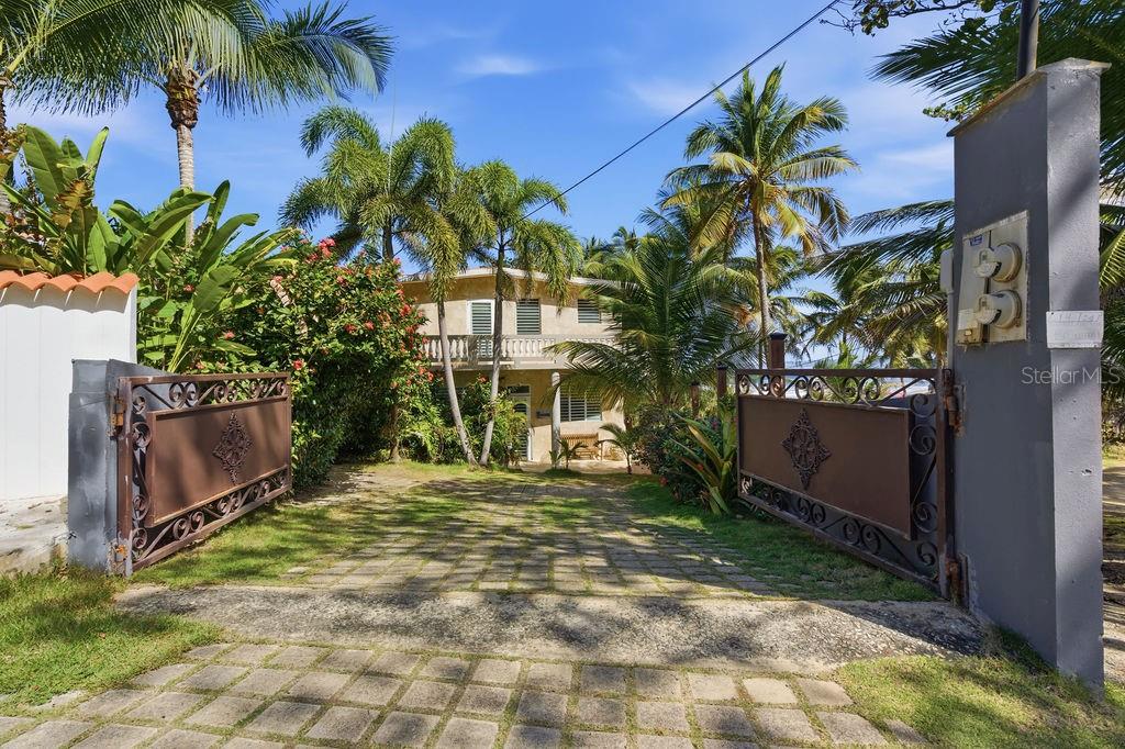 A paved driveway leads through an open gate toward a two-story house surrounded by palms — 26 BARRIO PUNTAS SANDY BEACH, RINCON, PR