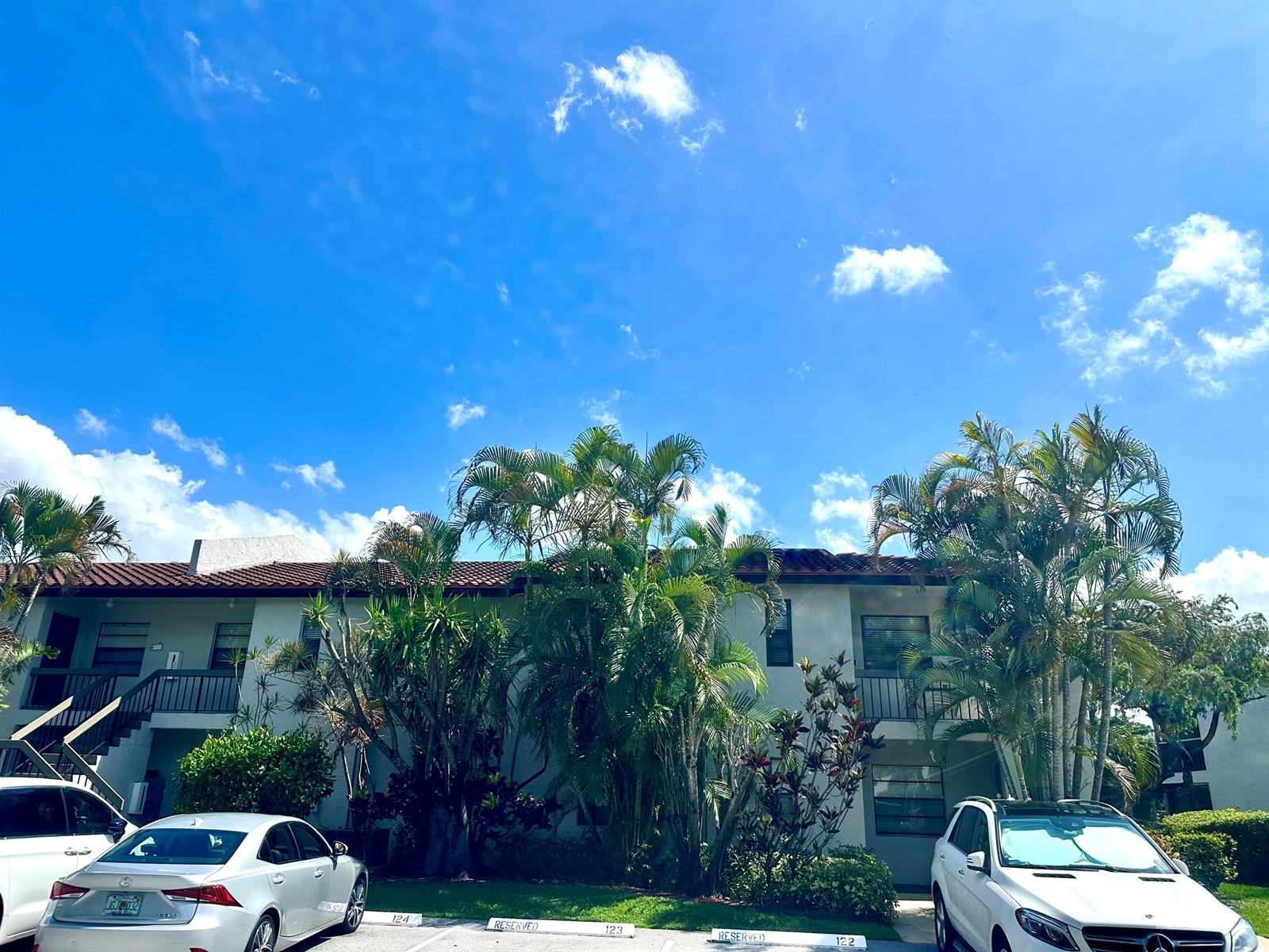 A two-story apartment building exterior features palm trees, reserved parking spaces, and a blue sky — 21535 Cypress Hammock Drive # 36j, Boca Raton FL 33428, BOCA RATON, FL