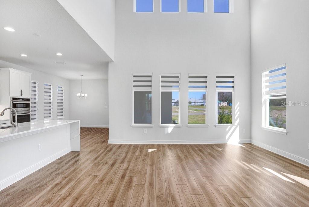 A bright, open-concept living area features light wood-look flooring, white walls, and tall windows — 28793 SWEET PERSIMMON DRIVE, WESLEY CHAPEL, FL