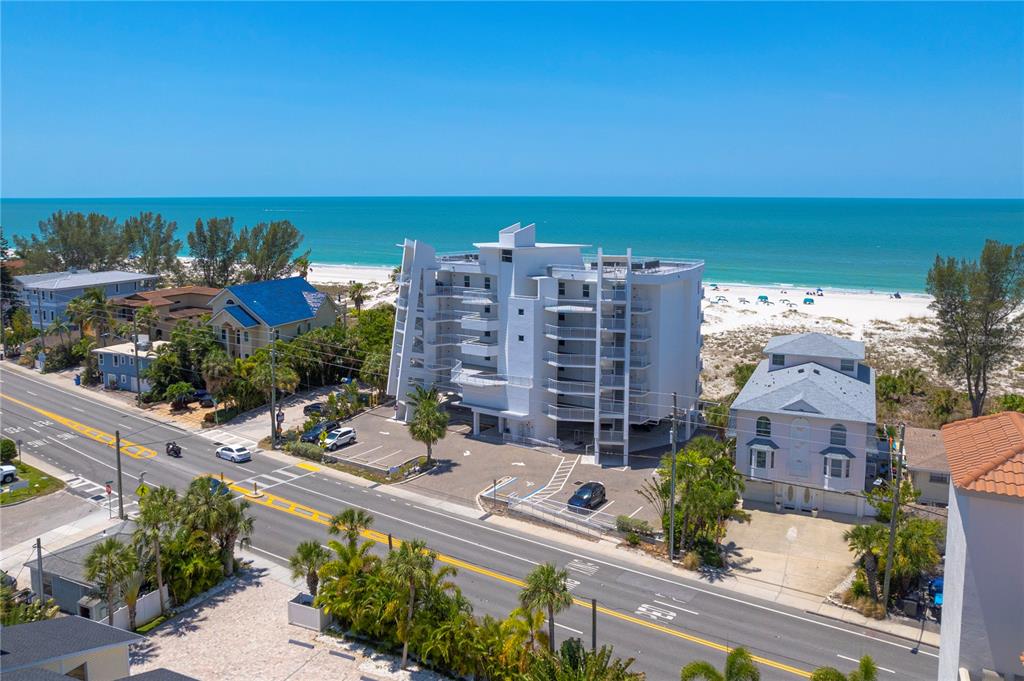 An aerial view shows a white multi-story condominium building located between a road and the beach — 10116 GULF BOULEVARD UNIT 403W, TREASURE ISLAND, FL
