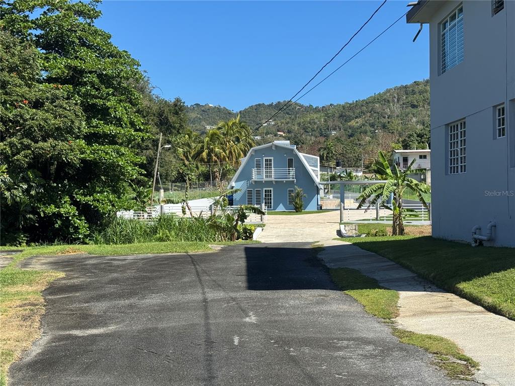 A blue barn-style house sits at the end of a paved driveway near mountains — 5 BO CELADA, GURABO, PR
