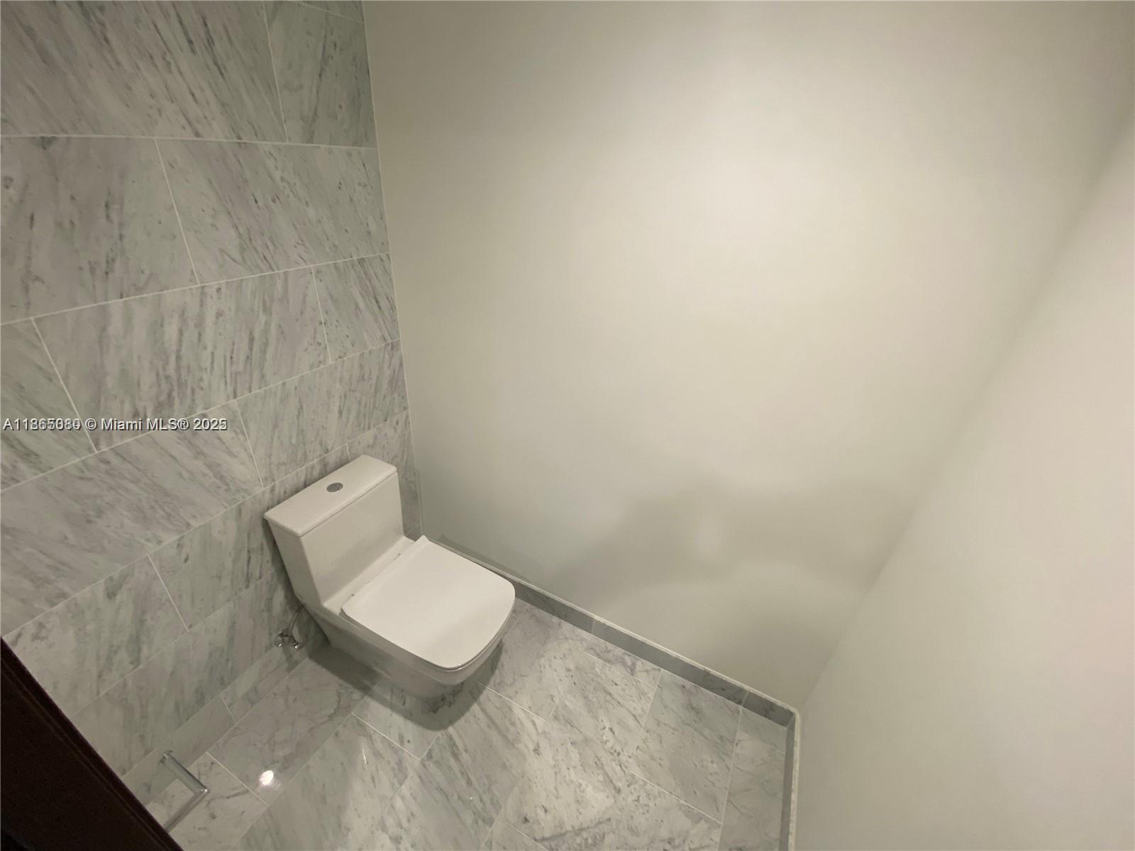 A modern bathroom features a white toilet, marble-tiled walls, and matching marble floor tiles — 1000 Brickell Plz # 3010, Miami FL 33131, MIAMI, FL