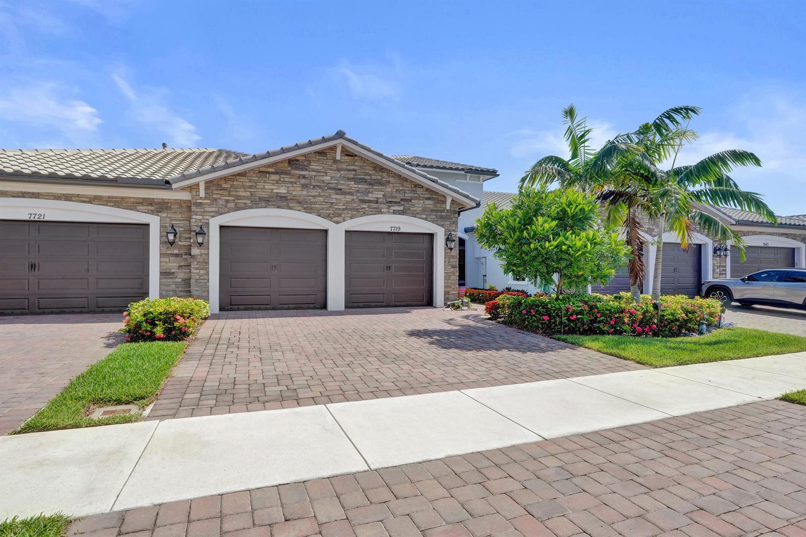 A stone-fronted house with a brick driveway and two garage doors under a blue sky — 7719 S Blue Spring Drive, Parkland FL 33067, PARKLAND, FL
