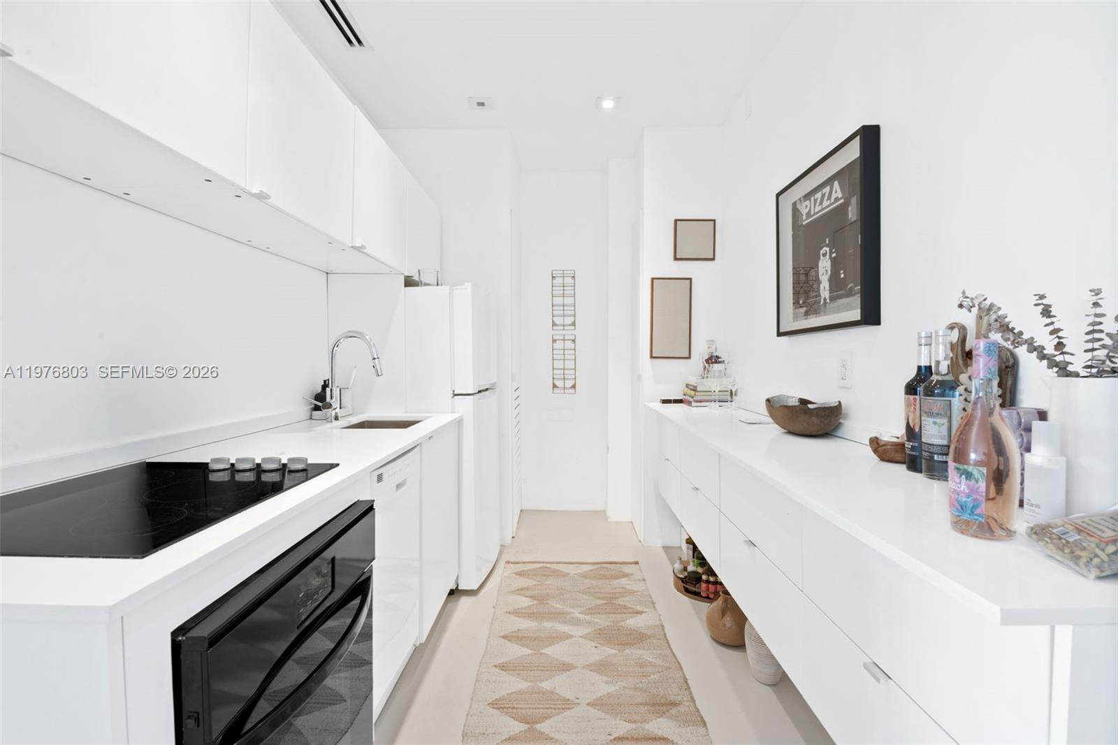 A narrow, all-white galley kitchen features minimalist cabinetry, a black cooktop, and a patterned rug — 3 Island Ave # G, Miami Beach FL 33139, MIAMI BEACH, FL